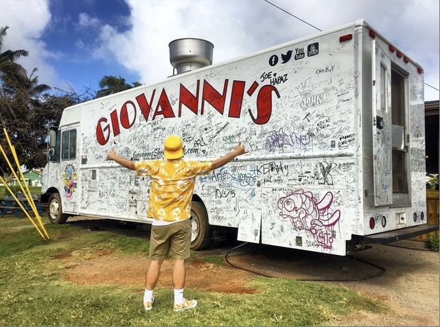 A visitor in a yellow aloha shirt and bucket hat stands with arms spread wide in front of the white Giovanni's shrimp truck in Kahuku, covered from end to end in guest signatures