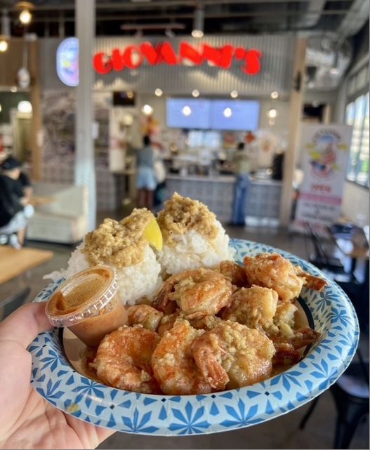 A Giovanni's shrimp plate held up in front of the Kakaʻako counter: garlic shrimp, two scoops of rice with garlic topping, a lemon wedge and a cup of sauce, with the red GIOVANNI'S sign and food hall blurred behind